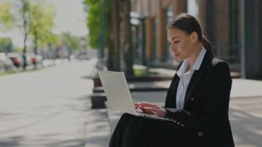 Tarz sahibi, çekici, koyu renk saçlı bir kadın ofis binasının yanında otururken kablosuz bilgisayarda yazı yazıyor. İnsan kavramı, teknoloji ve başarılı kariyer.