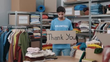 Kafkasyalı gönüllü adamın tabelalı portresi. Teşekkür cümlesiyle ve gülümseyerek kameraya bakıyor. Arkasında eşyalar olan raflar. Bağış kavramı