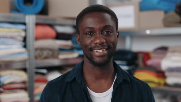 再利用服の中心で仕事をしながら広い笑顔でカメラを見ている多人種幸せ男のポートレート 第二の手と持続可能性の概念 — ストック動画