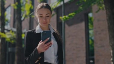 Kafkasyalı zarif iş kadını, modern akıllı telefondan e-postaları kontrol ederken iyi haberler alıyor. Kahverengi saçlı genç bayan ofis merkezinde yürürken mutluluktan el kol hareketi yapıyor..