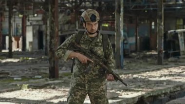 Kamuflaj giysileri ve miğfer takan kendine güvenen beyaz adam, elinde saldırı tüfeğiyle hasarlı çelik fabrikasında duruyor. Vatanseverlik ve savunucunun cesareti.