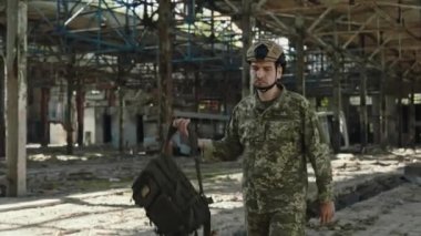 Kamuflaj giysileri giyen, sırt çantası taşıyan ve sanayi bölgesinde yürüyen cesur beyaz adam. Askeri asker savaş sırasında savunma görevinde..