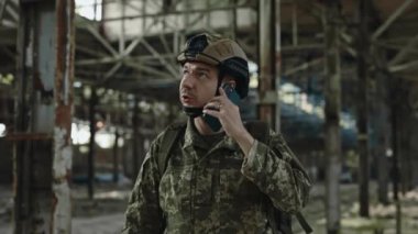 Ordu mensubu elinde akıllı telefonuyla bina bina üstüne bina yıkıp etrafa bakıyor. Miğferli ve üniformalı tatminkar ordu subayı mobil konuşma yapıyor..