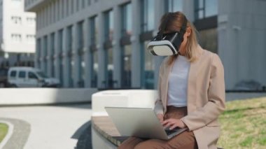 VR kulaklıklı beyaz kadın girişimci dizüstü bilgisayarla bankta oturuyor ve ofis binasının yakınında çalışıyor. İş, insan ve yeni teknoloji kavramı.