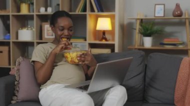 Afro-amerikan hamile anne dizüstü bilgisayardan favori programı izlerken sağlıksız atıştırmalıklar yiyor. Çekici hamile kadın evde zevkle yalnız başına dinleniyor..