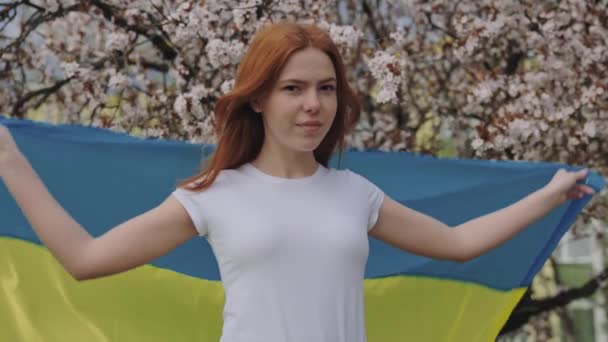 春天的樱桃园 美丽的红头发女人披着乌克兰国旗 微笑着看着镜头的高加索女性 并在战争期间呼吁支持乌克兰 — 图库视频影像