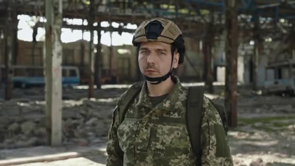 身穿军事迷彩服 头戴头盔的自信的高加索人站在被毁的钢铁厂上 目不转睛地看着相机 战争和入侵期间的军事力量 — 图库视频影像