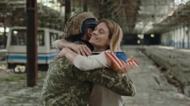 Amerikan erkek askerinin mutlu karısına sarıldığı ve Amerikan bayrağının elinde tahrip edilmiş bir imalat üzerinde durduğu görüntüler. Uzun zamandır beklenen savaş sonrası askeri aile toplantısı..