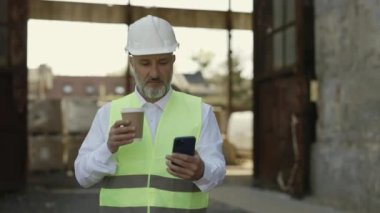 Kafkasyalı olgun ustabaşı modern akıllı telefonlarda internet taraması yapıyor ve inşaat alanında yürürken sıcak kahve içiyor. Miğferli sakallı mimar ve yansıtıcı yelek iş yerinde mola veriyor..