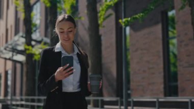 Successful smiling woman holding cardboard cup of takeaway coffee and using smartphone outdoors. Dark haired caucasian woman checking social networks while walking on city street.