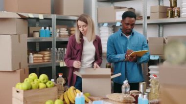 Depo çalışanlarından oluşan çok ırklı bir ekip yiyecek paketliyor ve not alıyor. Genç adam ve kadın günlük giysiler içinde markette teslimat için market hazırlıyorlar..