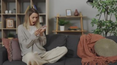 Depressed young woman with unwanted pregnancy looking on positive test while sitting with crossed legs on couch at home. Problems and changes in life.