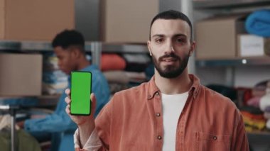 Beyaz adamın elinde akıllı telefon tutan, yeşil ekran görüntüsü olan ve depoda dikilirken kameraya gösteren portre görüntüsü. Arka plandaki çok ırklı meslektaşı.