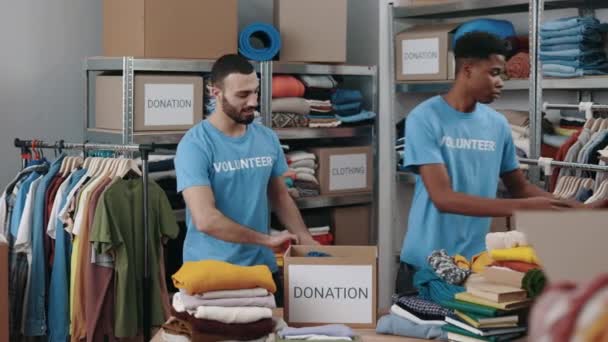 Two Male Volunteers Folding Clothes Putting Cupboard Box Warehouse Humanitarian — Video