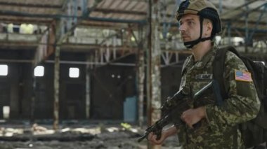 Terk edilmiş sanayi fabrikasında elinde tüfekle yürüyen Amerikan askerinin yan görüntüsü. Askerî üniformalı korkusuz bir subay düşmanla savaş sırasında savunmasını sürdürüyor..