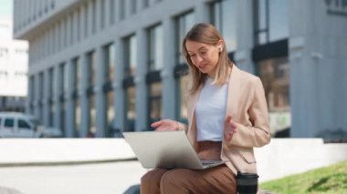 Smiling business woman talking to colleagues on laptop while sitting near office building on bench. Happy female manager in wireless earphones making video call conference outdoors.