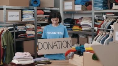 Portrait of energetic caucasian woman looking at the camera and smiling while holding banner with donation word in charity shop. Volunteer working in organization and sorting donated clothes