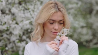 Sensual young blonde gently leaning blossoming branch to her face with spring garden on background. Relaxed woman looking at camera while spending time on fresh air.