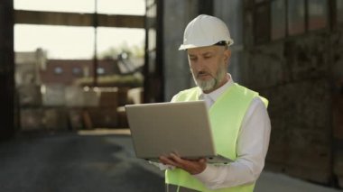 Kafkas mimarı modern dizüstü bilgisayarda doğru yapı hesaplaması için çalışıyor. Üniformalı gülümseyen olgun bir adam inşaat alanının önünde dikilip kameraya bakıyor..