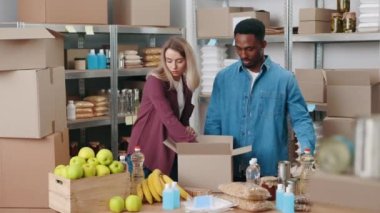 Çok kültürlü depo çalışanları günlük giysiler içinde karton kutuların içinde yiyecek paketliyorlar. Yakışıklı genç bir adam ve çekici bir kadın satış için gerekli düzenlemeleri yapıyor..