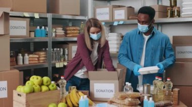 Afro-Amerikalı erkek ve beyaz kadın bağış paketleri hazırlamak için gıda bankasında birlikte çalışıyorlar. Kutuları toplarken yüz maskesi ve lastik eldiven takan gönüllüler..