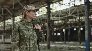 Caucasian female soldier walking through destroyed manufactory after war and looking around. Woman with backpack checking civil people presence on abandoned place.