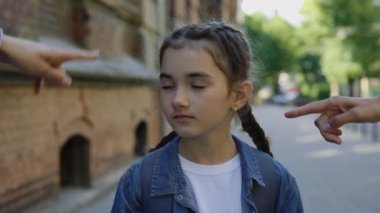 Close up of abusing little girl getting bullied outside near the school. Sad girl standing on the street and looking at camera. People pointing fingers schoolgirl