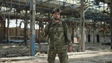 Elinde saldırı tüfeğiyle tahrip edilmiş çelik fabrikasında poz veren güçlü ve cesur bir adamın portresi. Kasklı ve üniformalı eğitimli erkek asker ciddi bir yüz ifadesiyle kameraya bakıyor..