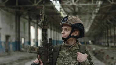 Confident female soldier with real weapon in hands standing among destroyed manufacture. Portrait of woman in military camouflage clothes looking at camera.
