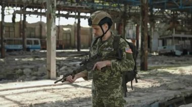 Amerikan ordusundan bir erkek subay terk edilmiş eski fabrikanın arasında elinde saldırı tüfeğiyle duruyor. Güvenlik kaskı ve üniformalı korkusuz bir adam yüzünde ciddiyetle kameraya bakıyor..