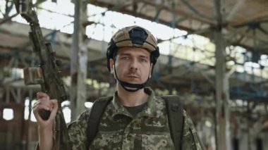 Portrait of soldier wearing military uniform, helmet and backpack looking at camera among ruined factory. Serious man holding black machine gun in his hand.