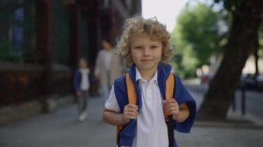 Little schoolboy standing on school yard holding bag turn around and looking at camera. Education concept