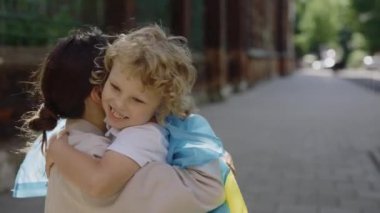 Little happy ukrainian child with ukrainian flag on shoulders hugs his mother on the street. near school. Stop war in Ukraine. Patriotic concept. Closeup