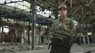 Patriotic soldier carrying backpack loaded with ammunition while walking on abandoned plant. Tired woman in uniform moving back home after completed military mission.