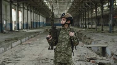 Askeri üniformalı, miğferli, tüfekli ve terk edilmiş bir fabrikanın önünde kameraya bakan cesur beyaz kadın. Savaş, savunma ve silah kavramı.