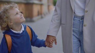 Son looking at his mother and holding her hand smilling on the road to school. Schoolboy with a backpack walking after lessons with his nanny. Education concept. Close up
