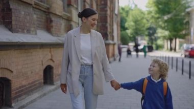 Mom and son are walking outside talking and smilling to each other. Happy family holding hands after the lessons in school were finished. Educational and people concept