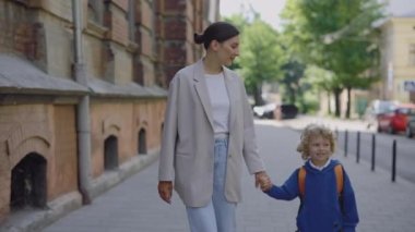 Young mother holding her son hands talking and leads child to school for a lesson in class. Mom and son walking outdoors schoolyard