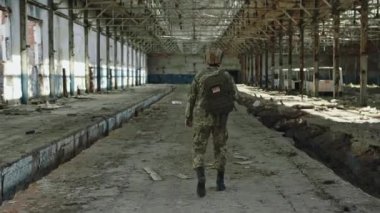Back view of male soldier from american forces walking with backpack. Patriot wearing protective helmet and camouflage uniform. Interior of a destroyed abandoned steel plant.