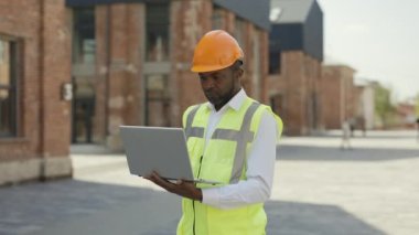 Smiling engineer in protective hardhat working with laptop looking at camera on construction site. Portrait of african american male architect checking project progress online.