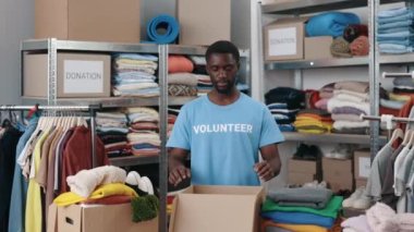 Portrait of male volunteer standing at the warehouse and looking at the camera while sorting and iterating clothes for donations. Belongings at the shelves at the background. Humanitarian aid concept