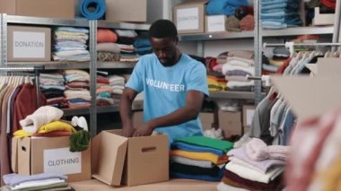 African american volunteer working at the warehouse while sorting and iterating clothes for donations. Belongings at the shelves at the background. Humanitarian aid concept
