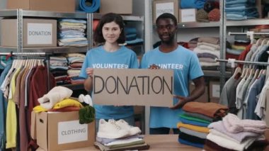 Portrait of energetic colleagues looking at the camera and smiling while holding banner with donation word in charity shop. Volunteers working in organization and sorting donated clothes