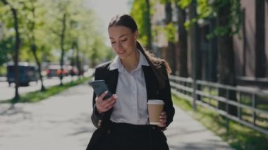 Charming business woman walking on city street with cup of coffee-to-go while and using modern smartphone. Smiling attractive entrepreneur taking coffee break outside business center.