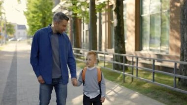 Close view of the father holds his sons hand, they leave school together and talk about how the day went. Father and son walking home after school day talking about lessons