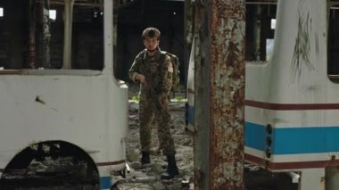 Concentrated soldier with sniper rifle standing between destroyed buses in abandoned plant. Military man wearing armour uniform maintaining territory of collapsed building.