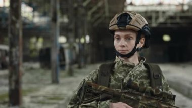 Serious fully equipped female soldier walking among captured building. Military woman with machine gun in hands patrolling large steel plant.