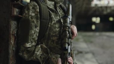 Side view of caucasian woman in camouflage military uniform and helmet standing with rifle on destroyed factory. Fear on face, enemy occupation.