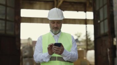 Senior male architect standing outside of building during construction process and typing message on modern smartphone. Positive man in reflective jacket and hardhat chatting online.