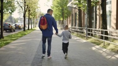 Back view of the father carrying backpack holding sons hand and son is holding the plant in a pot after ecology lesson at school and telling father stories from school or about his day
